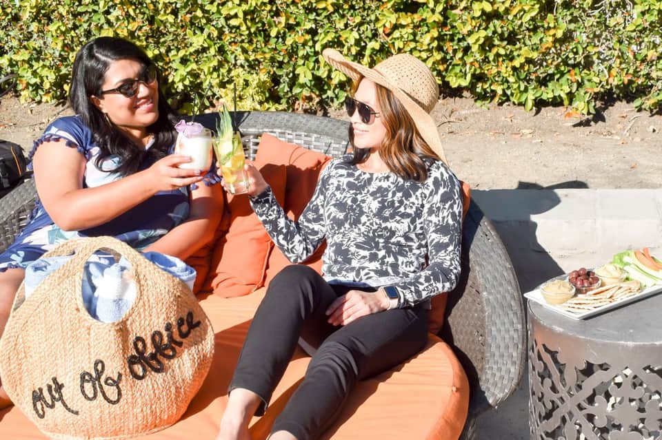 Two women enjoying drinks poolside with vineyard hedge backdrop