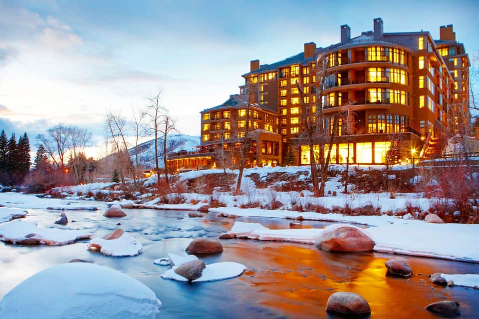 Luxury resort at dusk with glowing windows overlooking a partially frozen river surrounded by snow-covered landscape