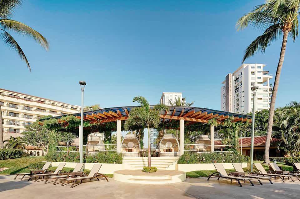 Resort courtyard with wooden pergola, lounge chairs, palm trees, and multi-story hotel buildings