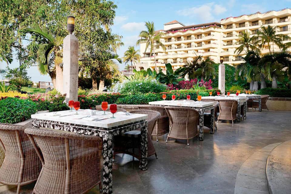 Outdoor dining terrace with wicker chairs, white tablecloths, red floral centerpieces, and resort buildings in background