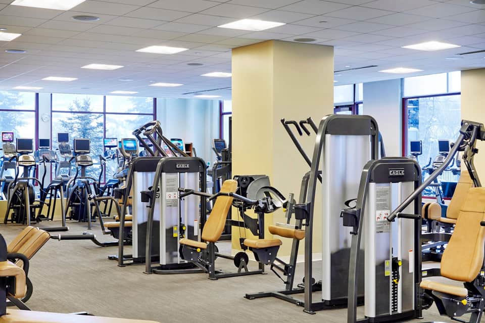 Modern gym facility with cardio equipment, exercise machines, and large windows overlooking trees