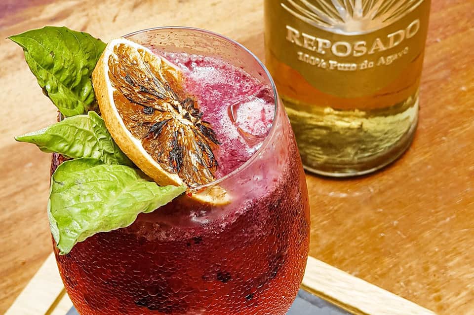 Tropical cocktail with pink liquid, dried citrus slice, and mint leaf next to Reposado bottle
