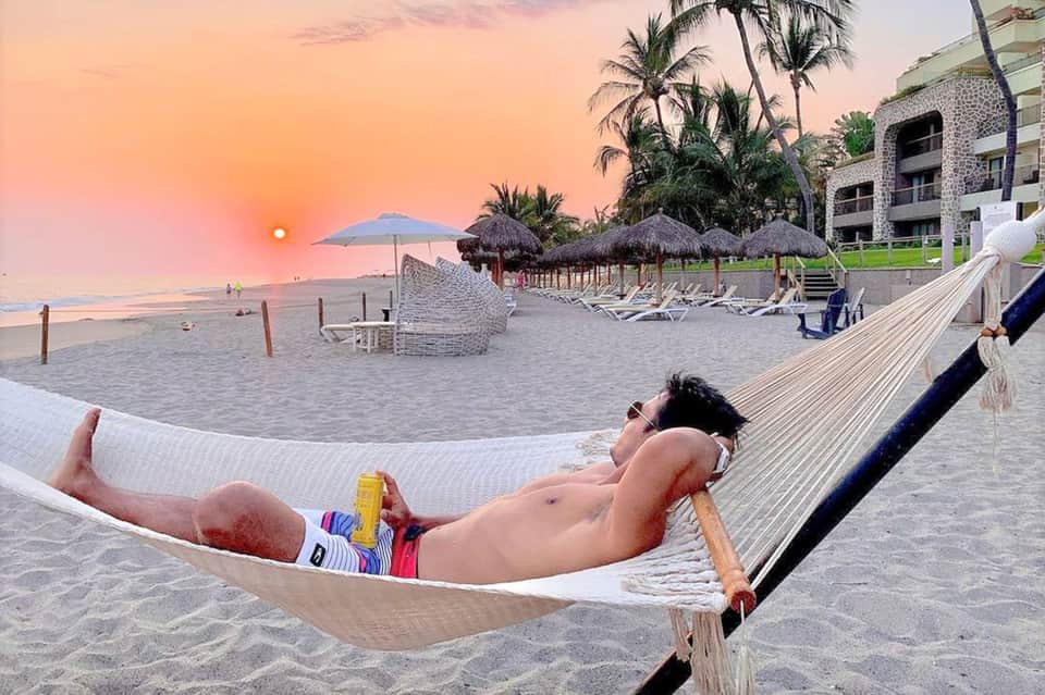 Person relaxing in hammock on beach at sunset with resort and umbrellas visible