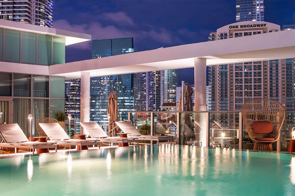 Rooftop pool at night with city skyline views, lounge chairs, and modern architecture