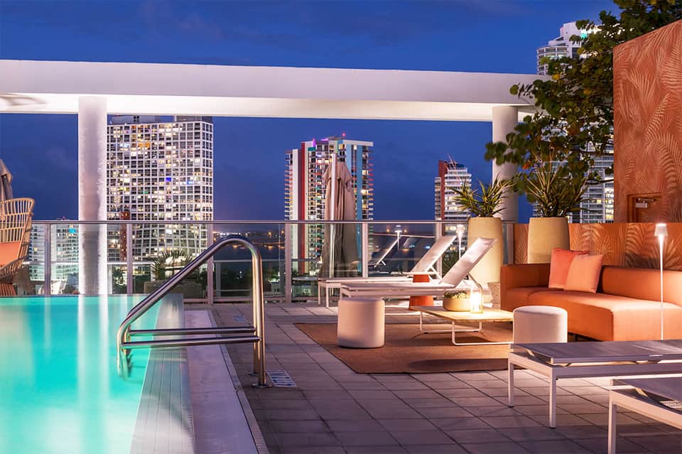 Illuminated rooftop pool and lounge area at night with city skyline views and orange seating