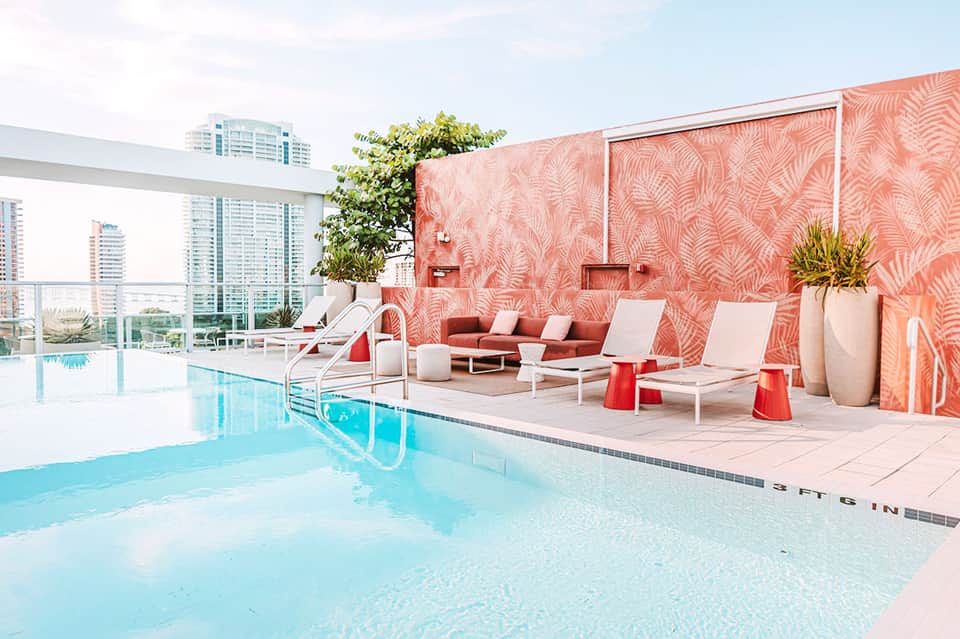 Indoor pool with coral patterned accent wall, white lounge chairs, and city skyline views through floor-to-ceiling windows