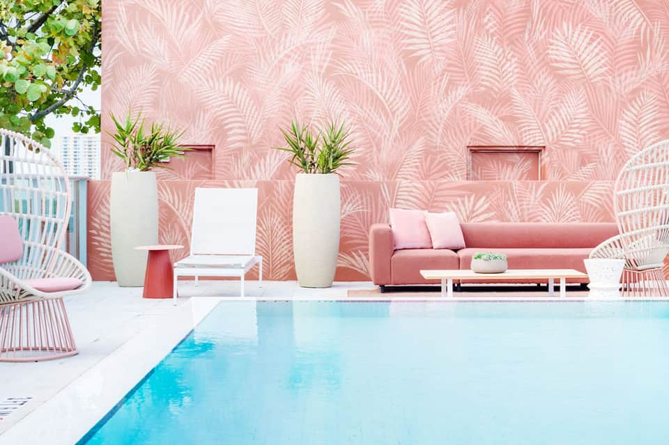 Modern poolside lounge with pink patterned wall, pink sofa, wicker chairs, and turquoise pool