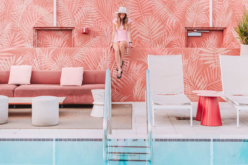 Woman in pink dress and white hat sitting on pool ladder in front of tropical patterned wall