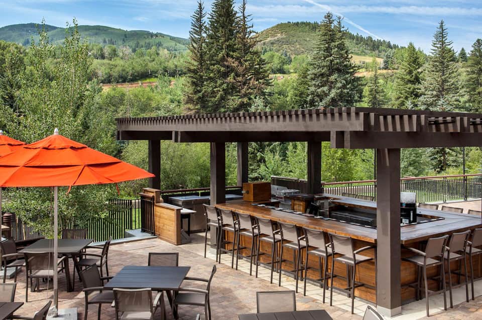 Outdoor mountain resort terrace with dark pergola, wooden bar, orange umbrella, and forested hillside views