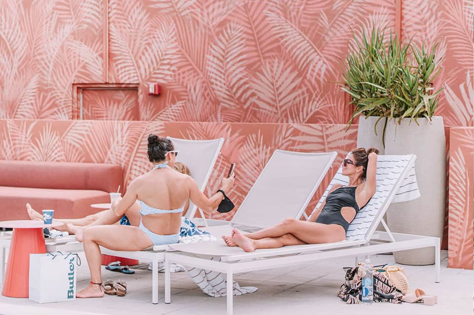Two women relaxing on lounge chairs against pink tropical leaf patterned wall