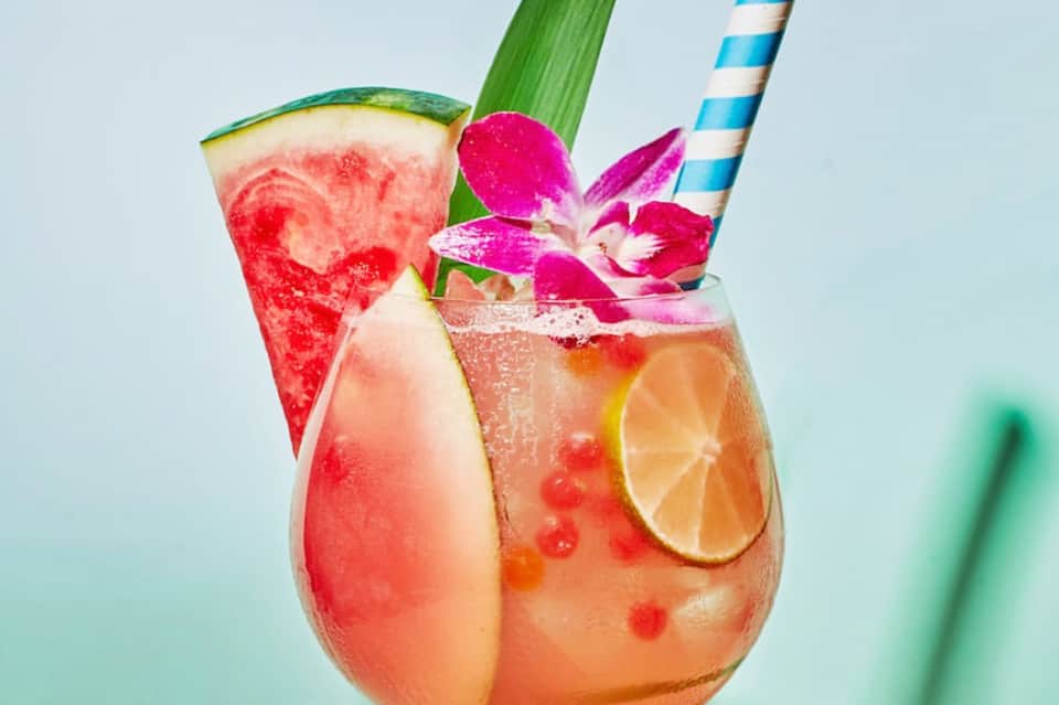 Tropical cocktail with watermelon slice, citrus, orchid flower and striped straws on light blue background