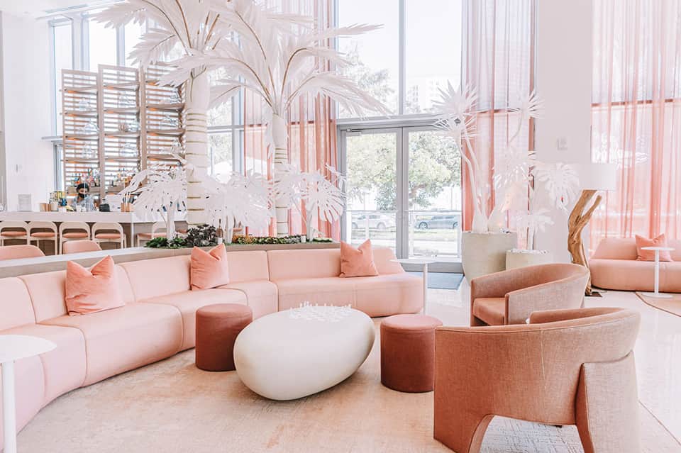 Contemporary lounge with curved pink sectional sofa, white ottomans, dried palm decor, and floor-to-ceiling windows