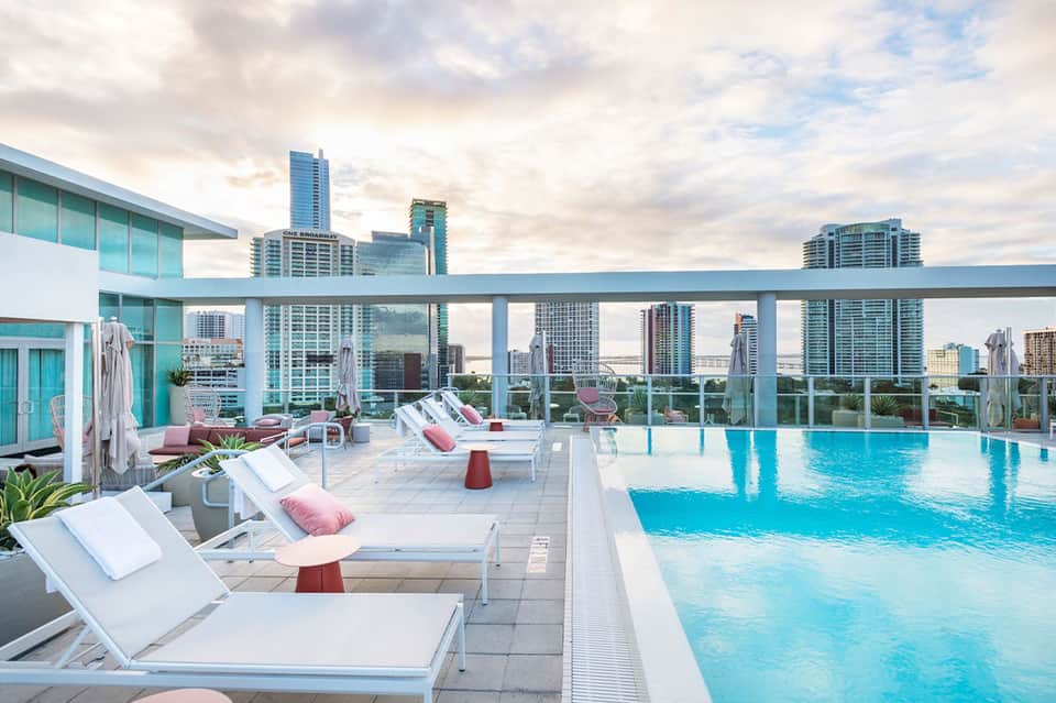 Resort rooftop pool with turquoise water, lounge chairs, and city skyline in background under cloudy sky