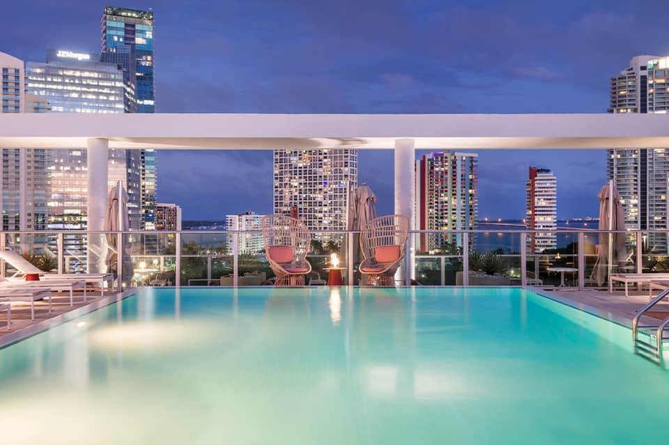 Rooftop infinity pool at dusk with city skyline, illuminated buildings, and modern lounge chairs