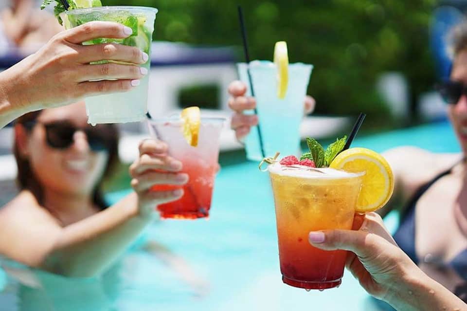 Colorful cocktails by pool with citrus garnishes and mint, guests in background