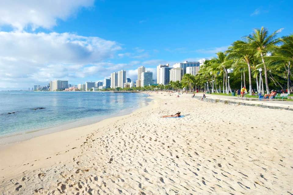 Waikiki Beach with white sand, palm trees, high-rise hotels, and visitors swimming and relaxing