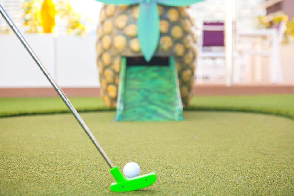 Mini golf course with green putter and white ball near a pineapple-themed obstacle
