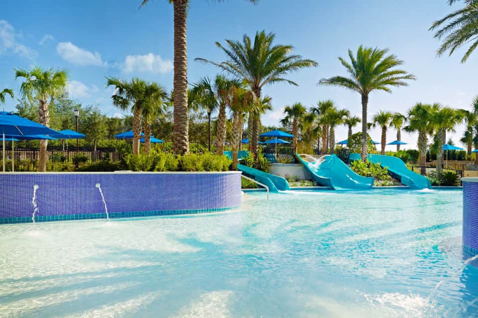 Resort pool with turquoise water, blue water slides, palm trees, and blue umbrellas under clear sky