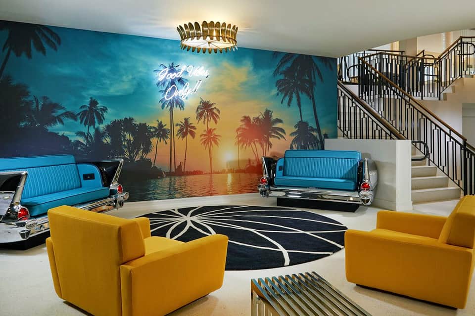 Retro-themed lobby with vintage car sculptures, palm mural, yellow chairs, and modern chandelier