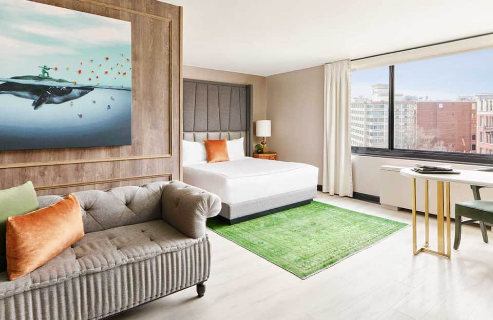 Luxury hotel suite with bed, sofa, whale artwork, green rug, city views, and modern furnishings