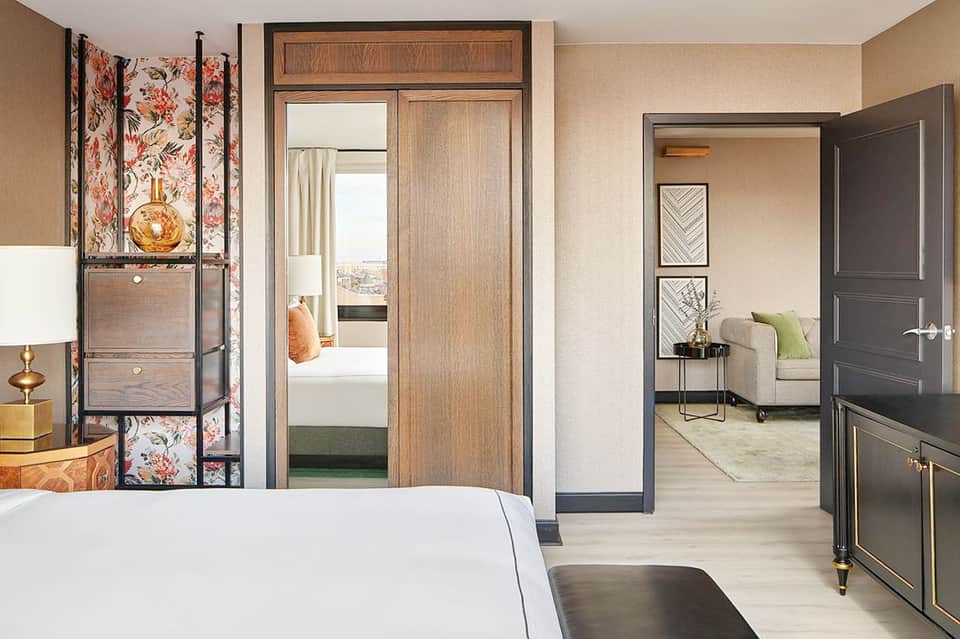 Spacious hotel suite with wooden doors, cream walls, and view into bedroom area