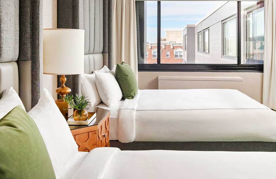 Modern hotel bedroom with two beds, green accent pillows, and city views