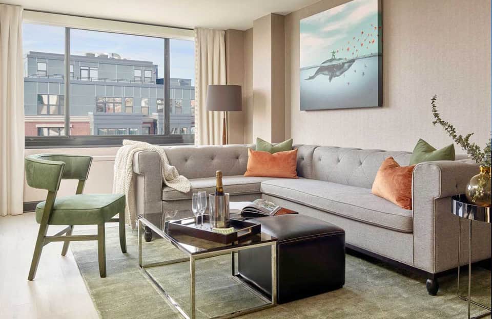 Modern living room with gray sectional sofa, orange and green pillows, large windows overlooking city