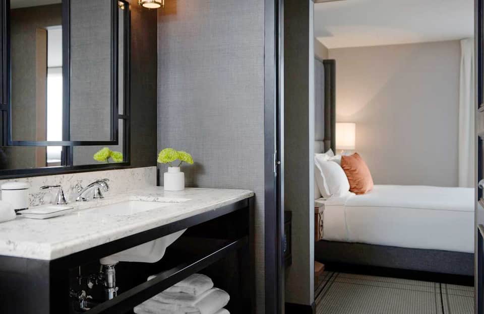 Modern hotel bathroom with marble countertop, black vanity, mirror, and view into bedroom with orange accent pillow
