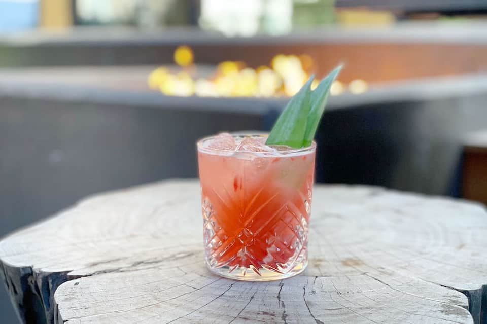 Coral-colored cocktail with ice and green leaf garnish served on wooden surface