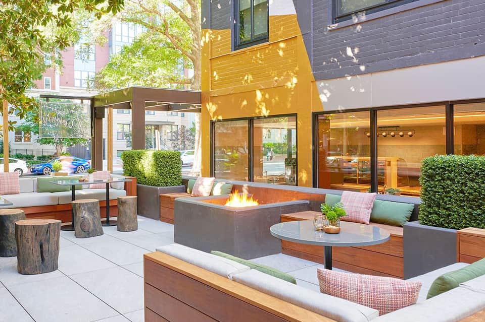 Urban rooftop lounge with fire pit, modern seating, yellow accent wall, and green landscaping