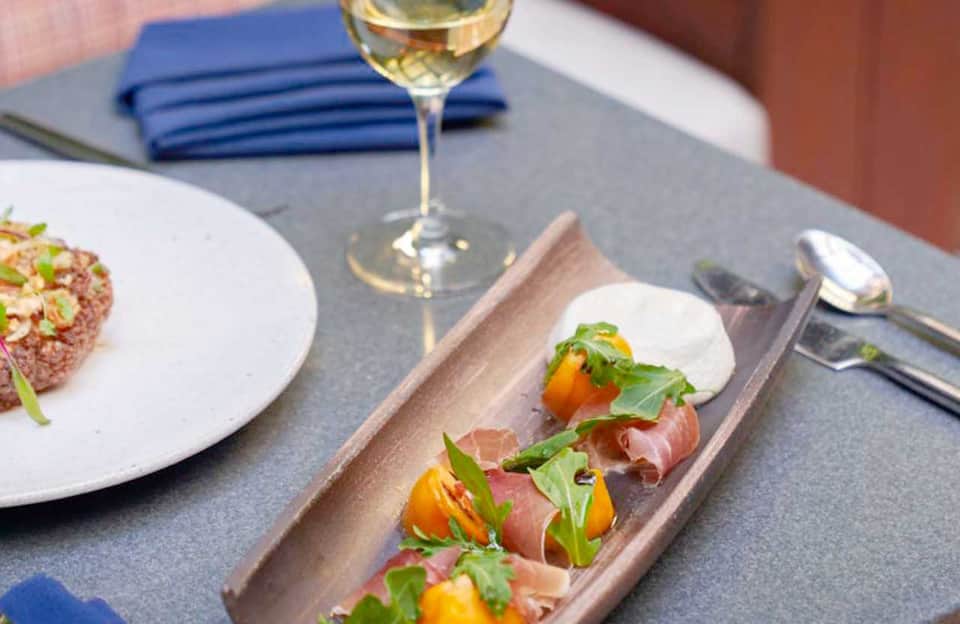 Fine dining plated appetizer with prosciutto, roasted peppers, and microgreens alongside white wine glass