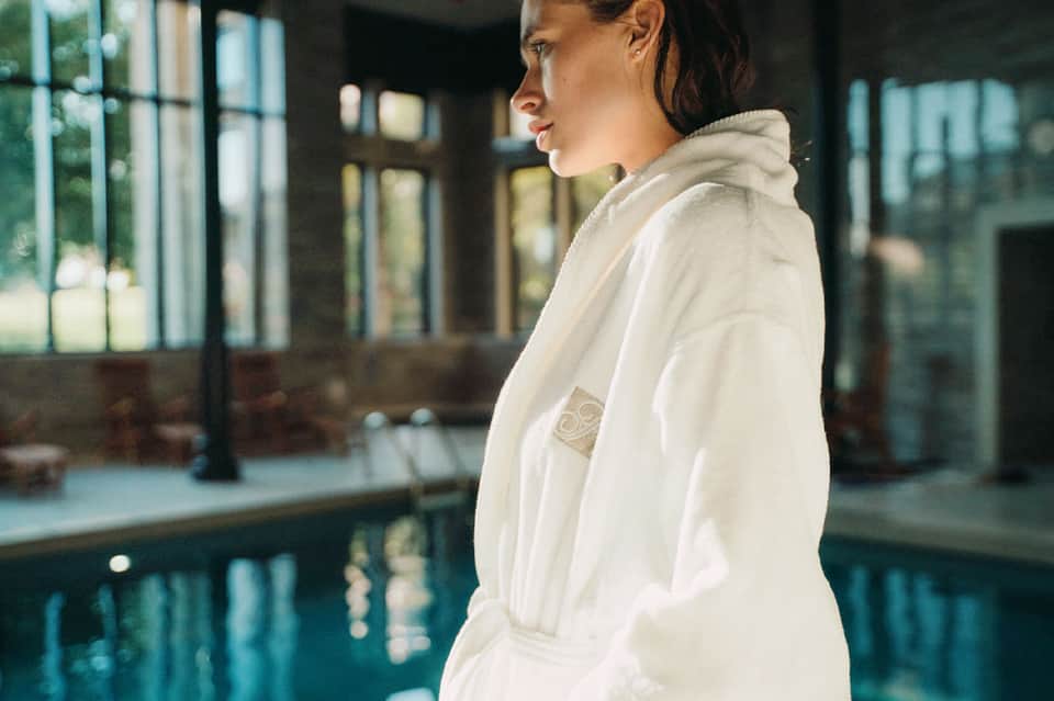 Woman in white spa robe standing by indoor swimming pool with modern architecture