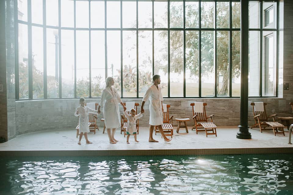 Family in white robes standing by indoor pool with large windows overlooking trees