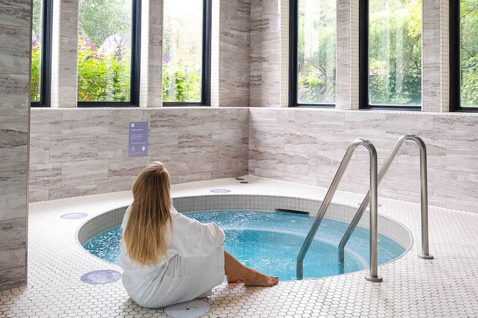 Woman in white robe sitting at edge of circular indoor hot tub with stone walls and large windows