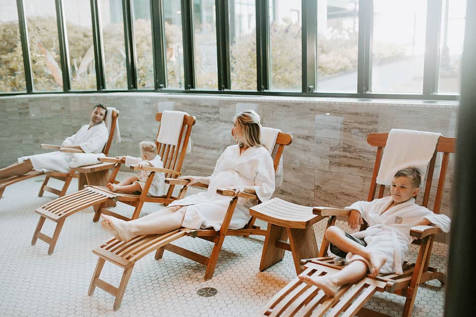 Spa relaxation room with guests in white robes sitting in wooden chairs overlooking water