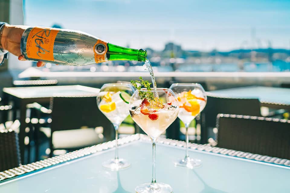 Champagne being poured into cocktail glasses with fruit garnish overlooking waterfront cityscape