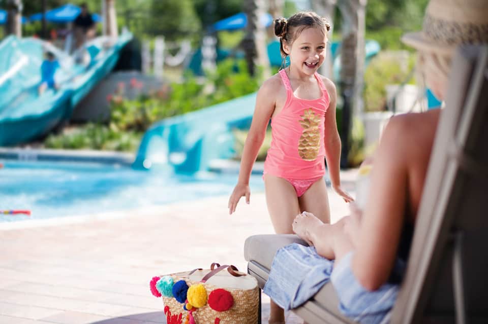 Young girl in pink swimsuit laughing with adult by pool area with water slides and lounge chairs