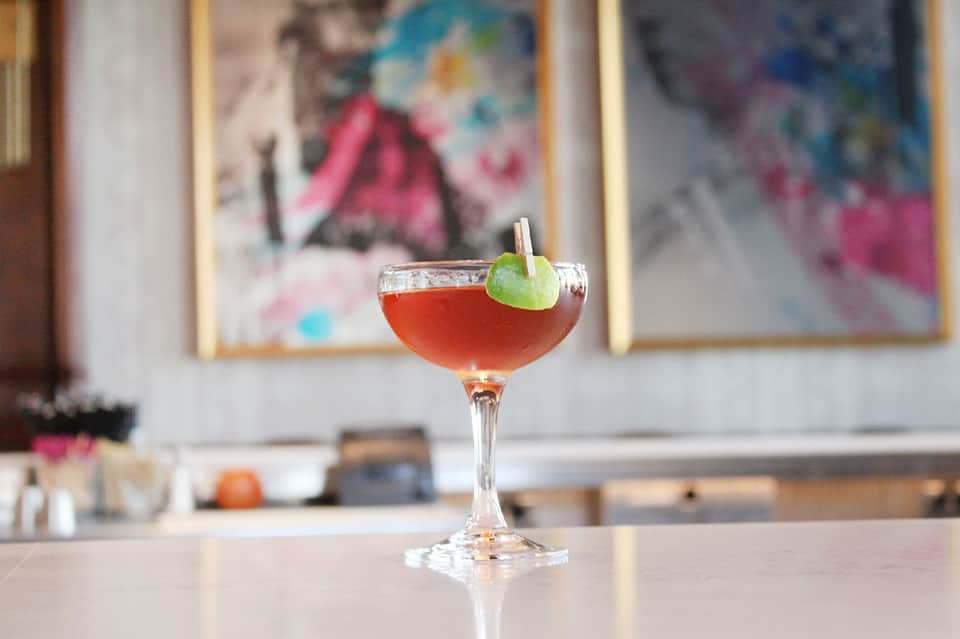 Craft cocktail with lime garnish on bar counter with blurred artwork background