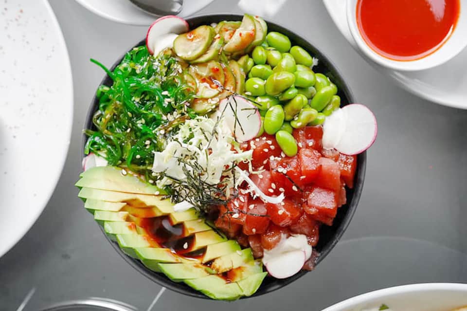 Colorful sushi bowl with seaweed, edamame, avocado, radish, salmon, and sesame seeds with red dipping sauce.
