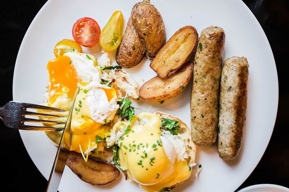 Breakfast plate with eggs Benedict, roasted potatoes, sausages, fresh tomato, and herbs on white plate