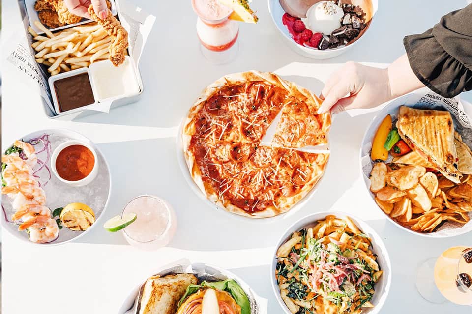 Overhead view of gourmet meal spread with pizza, seafood, drinks, and desserts