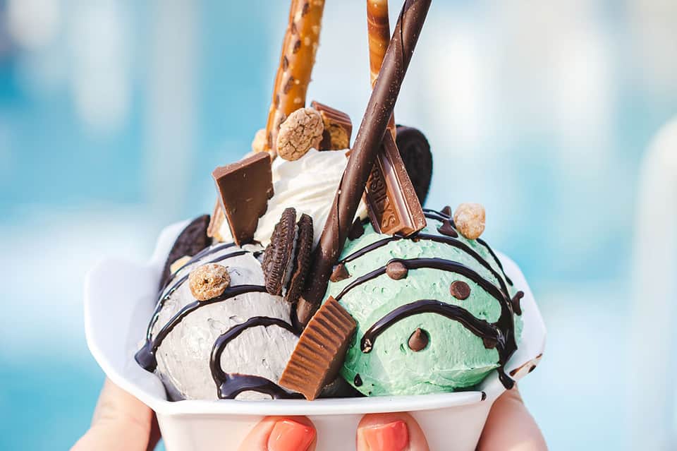 Colorful ice cream sundae with multiple flavors, chocolate sauce, and waffle cone held against blue sky