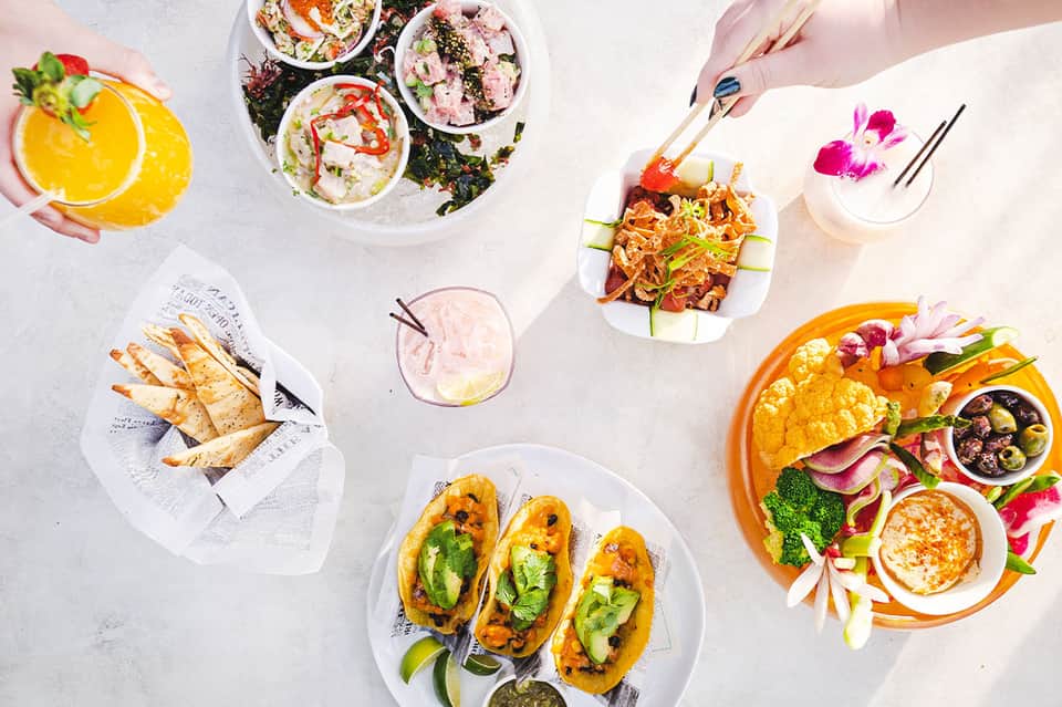 Overhead view of colorful plates of food, tropical drinks, and flowers on white table
