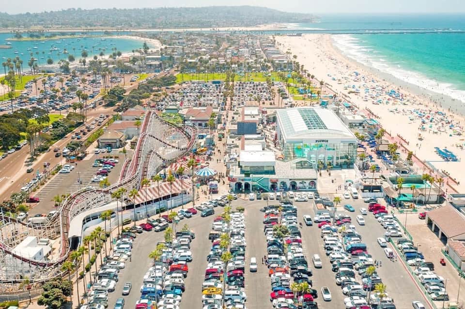 Aerial view of coastal amusement park with roller coaster, beach, parking lot, and boats in bay