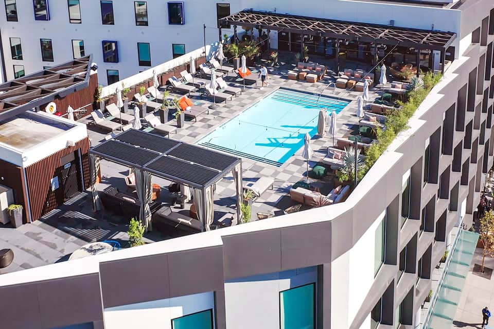Aerial view of a modern rooftop pool deck with lounge chairs, umbrellas, and surrounding hotel buildings