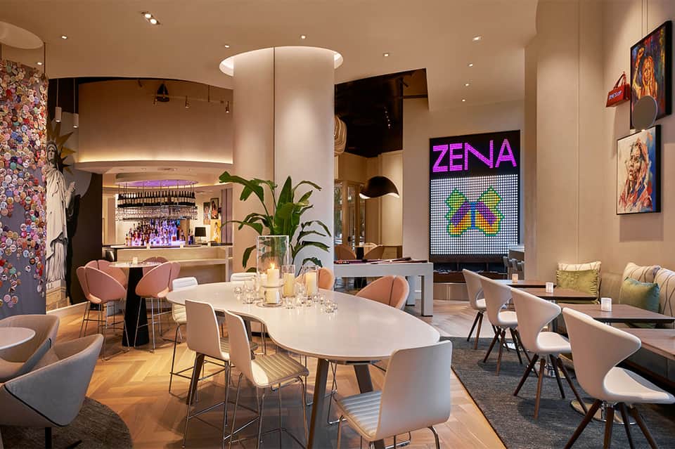Modern restaurant interior with white dining tables, pink chairs, bar area, and ZENIA neon sign
