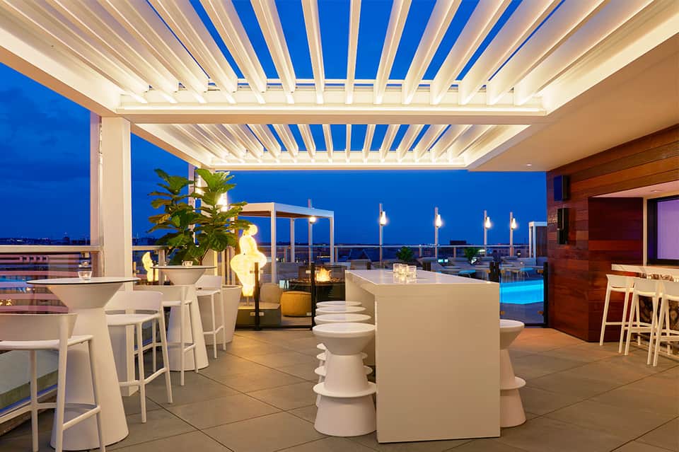 Modern rooftop bar with white lounge seating, pergola, evening blue sky, and city lights