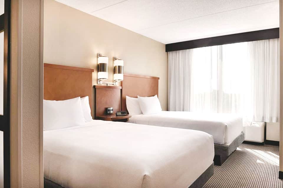 Hotel guest room with two queen beds, tan leather headboards, and large windows with sheer curtains