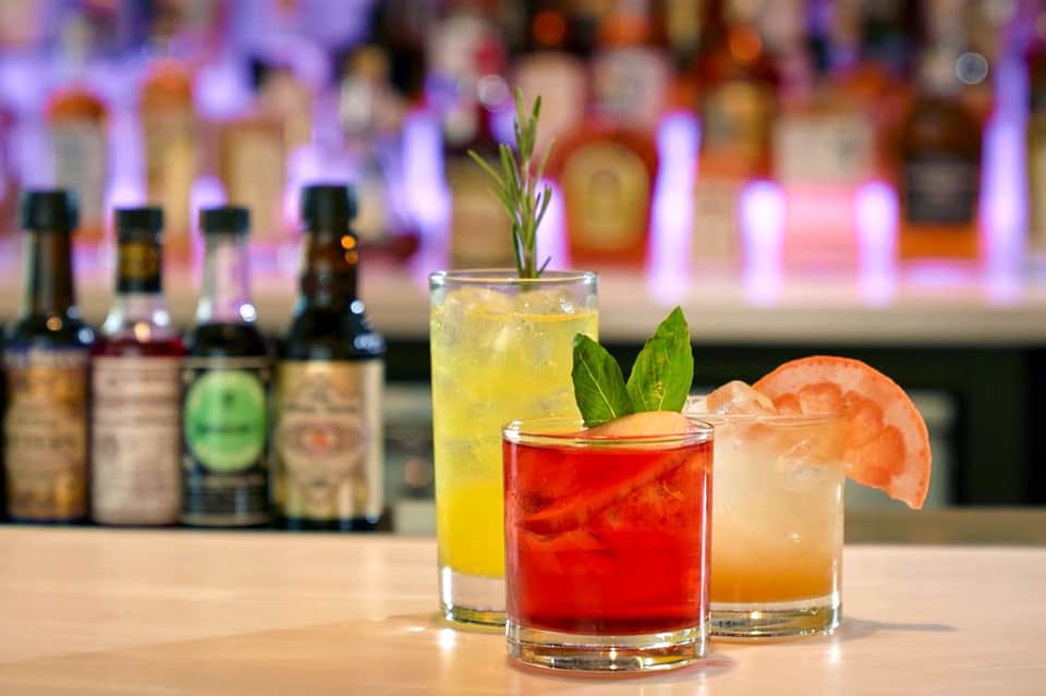 Three colorful cocktails on bar counter with bottles and purple lighting in background