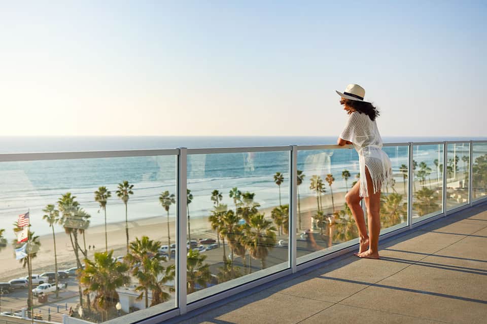 Woman in white cover-up and hat overlooking beach with palm trees from oceanview balcony